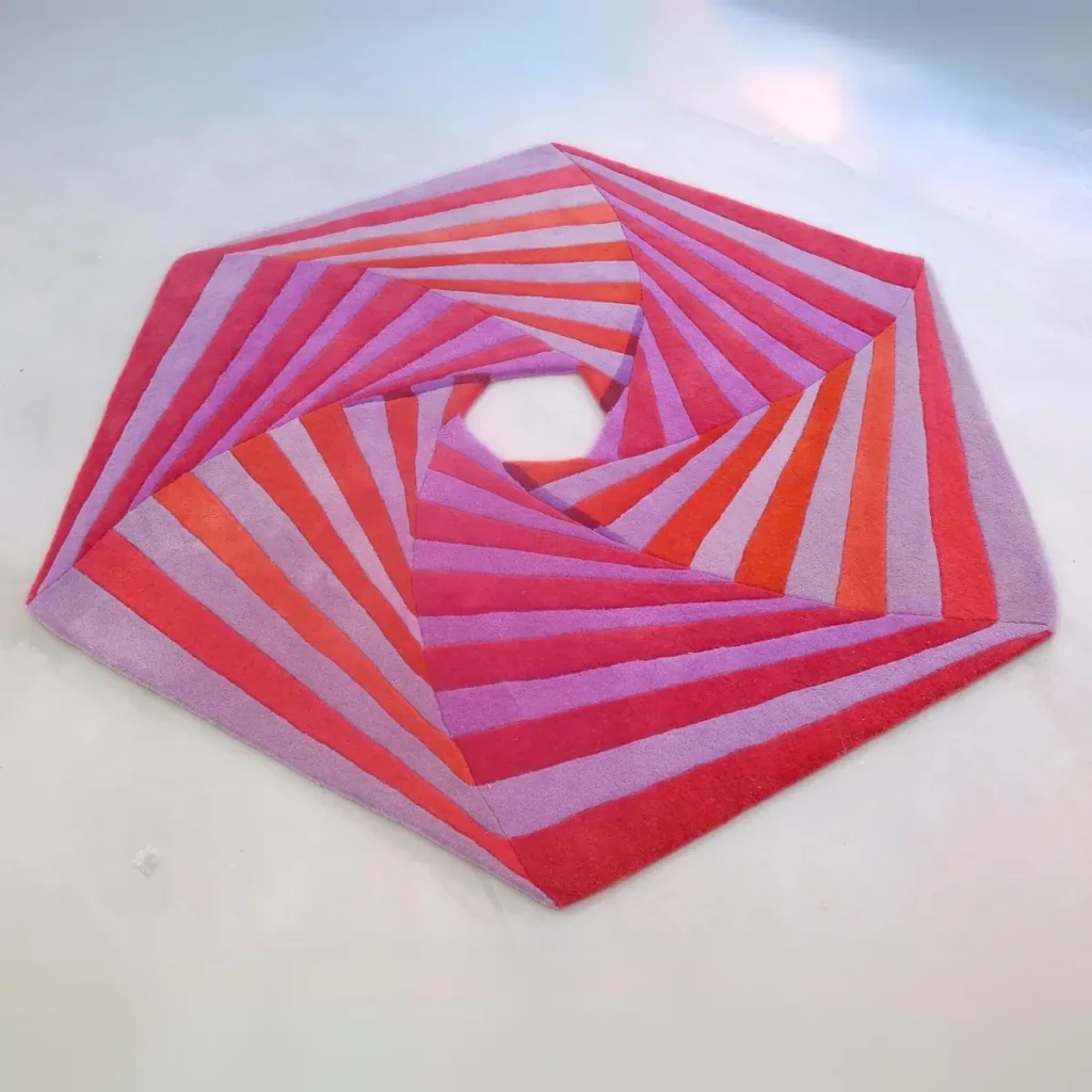 La peinture artistique « Swirl » d'Annika Johansson sous forme de tapis fait main rose et rouge. Tapis unique de forme hexagonale avec découpe au centre.